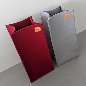 Op de afbeelding: Twee hoge opbergbakken van vilt. Eén is dieprood, de andere grijs. Elk heeft een leren patch met een logo. De bakken hebben een uniek, gebogen topontwerp en leunen tegen een witte muur.