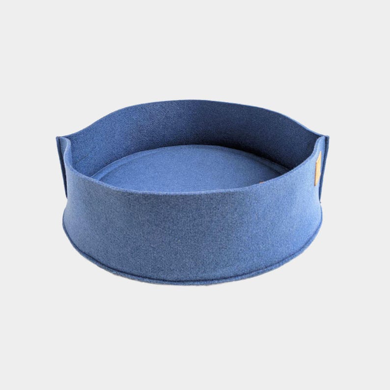 Peut inclure: Un lit pour animaux de compagnie rond, bleu, avec un bord sur&eacute;lev&eacute; et un int&eacute;rieur rembourr&eacute;. Fabriqu&eacute; en feutre, il comporte une petite &eacute;tiquette rectangulaire. Le design est simple et moderne, adapt&eacute; aux petits animaux.