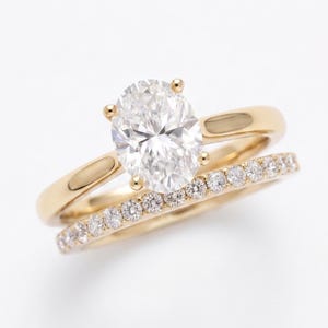 Puede incluir: Un conjunto de anillos de compromiso en oro amarillo con un diamante central de corte ovalado sujeto por cuatro garras en una banda lisa, junto a una alianza de boda a juego en oro amarillo con una hilera de pequeños diamantes redondos.