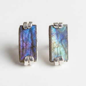 Puede incluir: Un par de pendientes de botón rectangulares. Cada pendiente presenta una piedra preciosa iridiscente pulida en tonos de azul, morado y verde, engastada en un marco de metal plateado. Diseño sencillo y moderno.