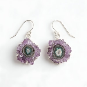 Peut inclure: Une paire de boucles d'oreilles pendantes composées de tranches d'améthyste brutes et circulaires, aux bords cristallins violets dentelés et au centre présentant des bandes blanches, grises et bleu sarcelle, montées sur des griffes en métal argenté.
