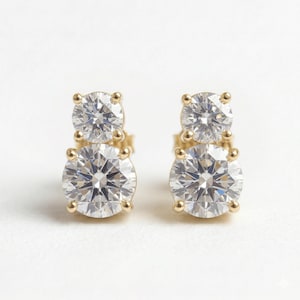 May include: A pair of stud earrings featuring two round, clear, faceted stones set vertically in yellow gold. Each earring consists of a smaller stone positioned above a larger stone, both secured by four-prong settings that hold the gems in place.