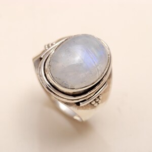 Pode incluir: Um anel de prata com um grande cabochão de pedra da lua oval e translúcido com um brilho azul iridescente. A pedra está cravada num bisel liso, rodeada por uma banda de prata em camadas com detalhes de contas decorativas nas laterais.