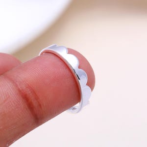 May include: A silver ring with a scalloped edge design. The ring is held by a finger, highlighting its shiny, reflective surface. The ring's design features a series of curved, wave-like shapes along its outer edge, creating a delicate, ornamental look.