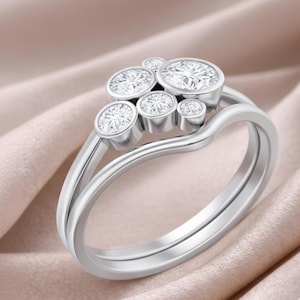 Puede incluir: Un anillo de plata con un grupo de siete piedras preciosas redondas y transparentes. El anillo tiene un diseño de doble banda, con una banda superior curvada. Las piedras están engastadas en biseles individuales, creando un efecto brillante. El anillo se muestra sobre una tela suave color melocotón.