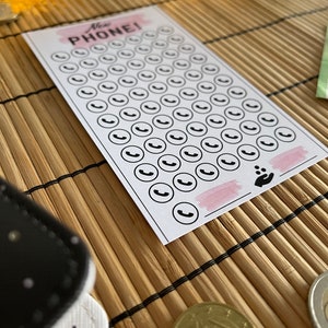 May include: A white card with the text "New Phone!" printed in black. The card has 50 black phone icons printed on it. The card is on a brown bamboo mat.