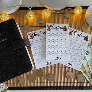 Könnte beinhalten: Drei Weihnachts-Spartabellen mit einem festlichen Rentier-Design. Jede Tabelle hat 25 Kreise, um Ersparnisse mit Beträgen von 10 Euro, 25 Euro und 50 Euro zu verfolgen. Die Tabellen sind mit dem Gesamtsparbetrag von 200 Euro, 250 Euro und 500 Euro beschriftet.