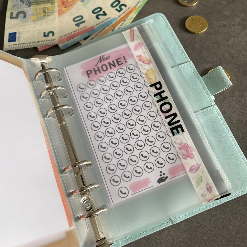 May include: A light blue leather bound planner with a clear plastic pocket on the front. The pocket contains a sheet of paper with the text "New Phone!" and a grid of phone icons. The pocket is labeled "PHONE".