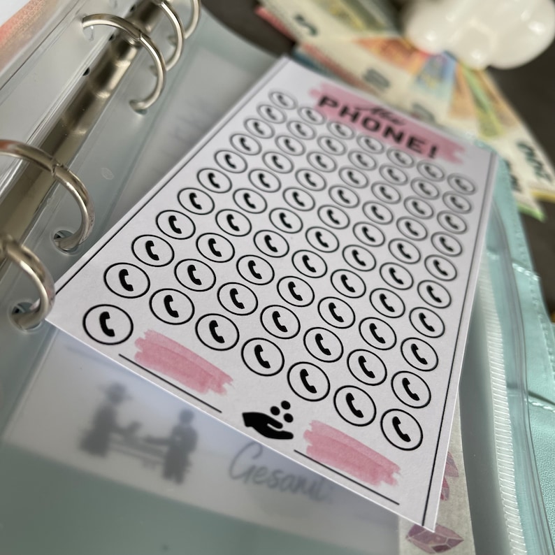 May include: A white sheet of paper with the text "The Phone!" printed in black. The paper is filled with 64 black phone icons in white circles. There is a pink watercolor wash at the bottom of the sheet.