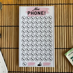 May include: A printable checklist with the text "New Phone!" at the top. The checklist has 70 phone icons in a grid pattern. There is a pink watercolor background behind the text and a pink watercolor line at the bottom of the checklist.