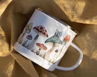 May include: A white enamel mug featuring a colorful illustration of various mushrooms and a small snail. The mug has a silver-toned metal rim and a matching white handle, showcasing a detailed botanical-style print with earthy tones.