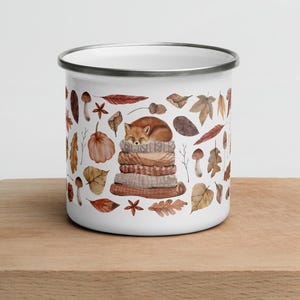 White enamel mug with a silver rim featuring a cute illustrated fox sleeping on a stack of cozy blankets surrounded by autumn leaves, pumpkins, mushrooms, and fall elements, displayed on a wooden board against a light background.
