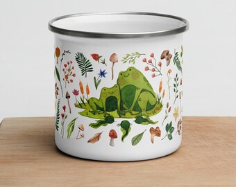 May include: A white enamel mug featuring a colorful illustration of two green frogs sitting together, surrounded by various small botanical elements including mushrooms, ferns, wildflowers, and leaves, with a silver-toned metal rim.