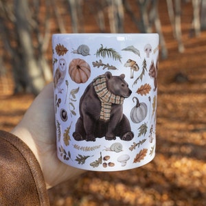 May include: White ceramic mug featuring a brown bear wearing a plaid scarf, surrounded by autumn-themed illustrations. The design includes pumpkins, leaves, mushrooms, and an owl. The mug is held against a blurred outdoor background.