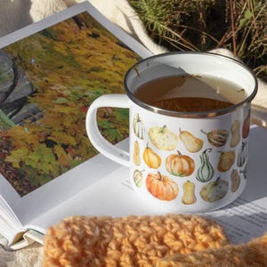 Puede incluir: Una taza de esmalte blanco con borde plateado, llena de un l&iacute;quido claro. La taza presenta un patr&oacute;n repetitivo de estilo acuarela de varias calabazas naranjas, amarillas y verdes de diferentes formas y tama&ntilde;os impresas en el exterior.