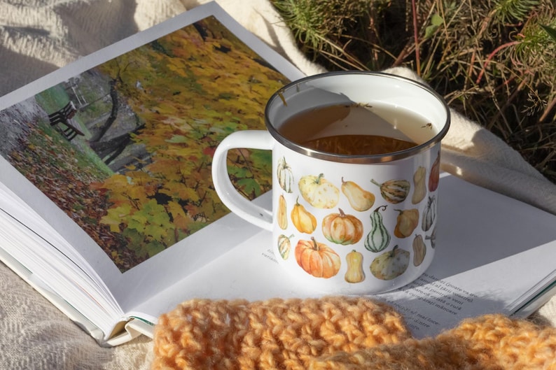 Puede incluir: Una taza de esmalte blanco con borde plateado, llena de un l&iacute;quido claro. La taza presenta un patr&oacute;n repetitivo de estilo acuarela de varias calabazas naranjas, amarillas y verdes de diferentes formas y tama&ntilde;os impresas en el exterior.
