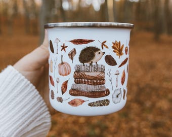 May include: A white enamel mug with a silver rim featuring a colorful illustration of a small hedgehog perched on a stack of folded sweaters, surrounded by various autumn elements including pumpkins, mushrooms, acorns, and fallen leaves.