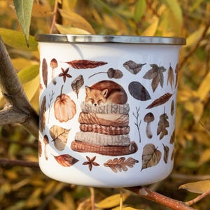 May include: A white enamel mug with a silver rim featuring a printed illustration of a fox sleeping on a stack of folded sweaters, surrounded by various autumn leaves, mushrooms, acorns, and star anise in shades of brown, orange, and tan.