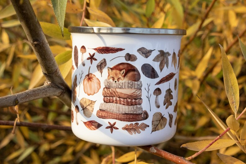 May include: A white enamel mug with a silver rim featuring a printed illustration of a fox sleeping on a stack of folded sweaters, surrounded by various autumn leaves, mushrooms, acorns, and star anise in shades of brown, orange, and tan.