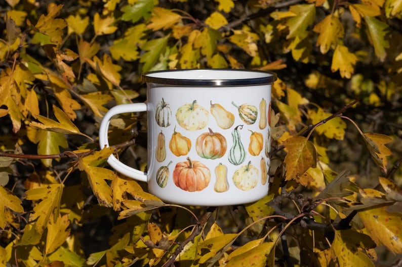Puede incluir: Una taza de esmalte blanco con un patr&oacute;n colorido de varias calabazas en tonos naranja, amarillo y verde. La taza tiene un acabado blanco liso, un borde de metal plateado y un asa blanca resistente situada en el lado izquierdo.