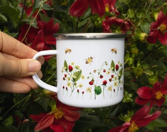 May include: A white enamel mug with a silver rim featuring a botanical illustration of wild strawberries, green leaves, small white flowers, and three flying honeybees. The mug has a white handle and a smooth, cylindrical body design.