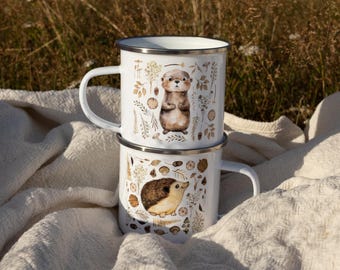 May include: Two white enamel mugs stacked vertically. The top mug features a brown otter illustration surrounded by small botanical elements. The bottom mug displays a brown hedgehog illustration with similar botanical motifs. Both mugs have silver rims.