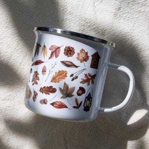 May include: A white enamel mug with a silver rim and a handle, featuring a repeating pattern of brown and orange autumn leaves, acorns, pinecones, a small wooden birdhouse, and a vintage-style lantern printed on the exterior surface.