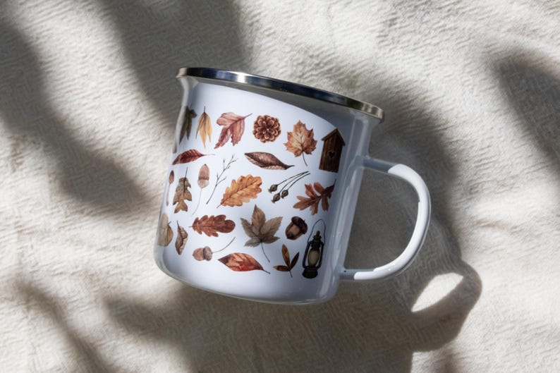 May include: A white enamel mug with a silver rim and a handle, featuring a repeating pattern of brown and orange autumn leaves, acorns, pinecones, a small wooden birdhouse, and a vintage-style lantern printed on the exterior surface.