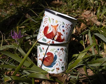 May include: Two white enamel mugs stacked vertically, featuring colorful illustrations of foxes and an otter surrounded by various wildflowers, mushrooms, and greenery. Each mug has a smooth white finish with a metallic silver rim at the top edge.