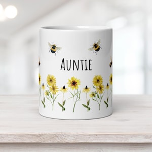 May include: A white ceramic mug featuring a printed design of yellow wildflowers and bumblebees. The word AUNTIE is printed in black, sans-serif capital letters in the center of the mug above the floral pattern that wraps around the base.
