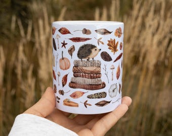 May include: A white ceramic mug featuring a repeating autumnal pattern of a small hedgehog perched on a stack of folded sweaters, surrounded by illustrations of orange pumpkins, brown fallen leaves, acorns, mushrooms, and slices of pie.