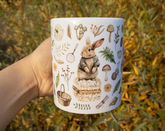 May include: A white ceramic mug featuring a watercolor-style illustration of a brown rabbit wearing a jacket, standing on a stack of books. The design includes various nature-themed elements like mushrooms, ferns, a magnifying glass, and a basket.