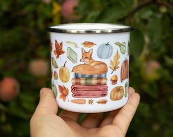 May include: A white enamel mug with a silver rim featuring a watercolor illustration of a fox resting on a stack of folded blankets. The design includes autumn leaves, pumpkins, a book, a sweater, and various seasonal harvest elements.