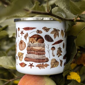 May include: A white enamel mug with a silver rim featuring a watercolor illustration of a sleeping fox curled up on a stack of folded sweaters, surrounded by various autumn leaves, acorns, mushrooms, and star anise in shades of brown and orange.