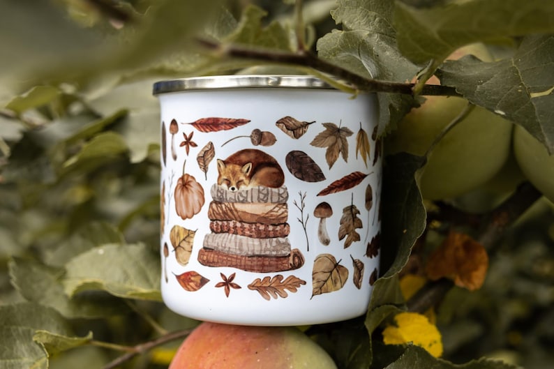 May include: A white enamel mug with a silver rim featuring a watercolor illustration of a sleeping fox curled up on a stack of folded sweaters, surrounded by various autumn leaves, acorns, mushrooms, and star anise in shades of brown and orange.