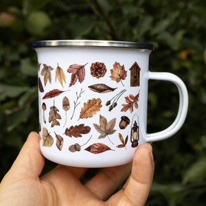 May include: A white enamel mug with a silver rim and a handle, featuring a repeating pattern of brown and orange autumn leaves, acorns, a pinecone, a small wooden birdhouse, and a vintage-style lantern printed on the exterior surface.
