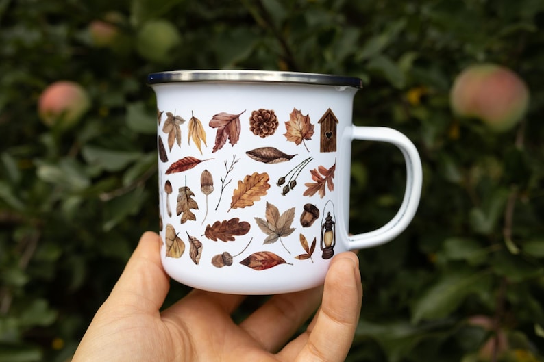 May include: A white enamel mug with a silver rim and a handle, featuring a repeating pattern of brown and orange autumn leaves, acorns, a pinecone, a small wooden birdhouse, and a vintage-style lantern printed on the exterior surface.