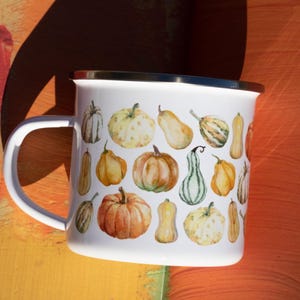 Puede incluir: Una taza de esmalte blanco con un colorido patr&oacute;n de estilo acuarela de varias calabazas y calabacines en tonos naranja, amarillo, verde y crema. La taza tiene un acabado blanco liso, un asa curva y un borde superior de metal plateado.