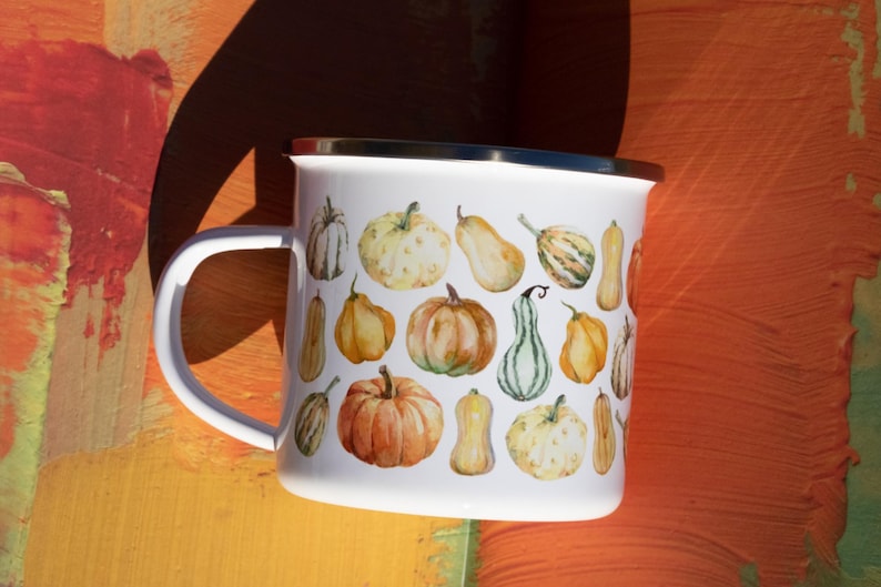Puede incluir: Una taza de esmalte blanco con un colorido patr&oacute;n de estilo acuarela de varias calabazas y calabacines en tonos naranja, amarillo, verde y crema. La taza tiene un acabado blanco liso, un asa curva y un borde superior de metal plateado.