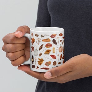 Op de afbeelding: Witte keramische mok met een herfstthema. De mok is bedekt met een patroon van herfstbladeren, pompoenen, eikels, paddenstoelen en andere herfstachtige afbeeldingen.