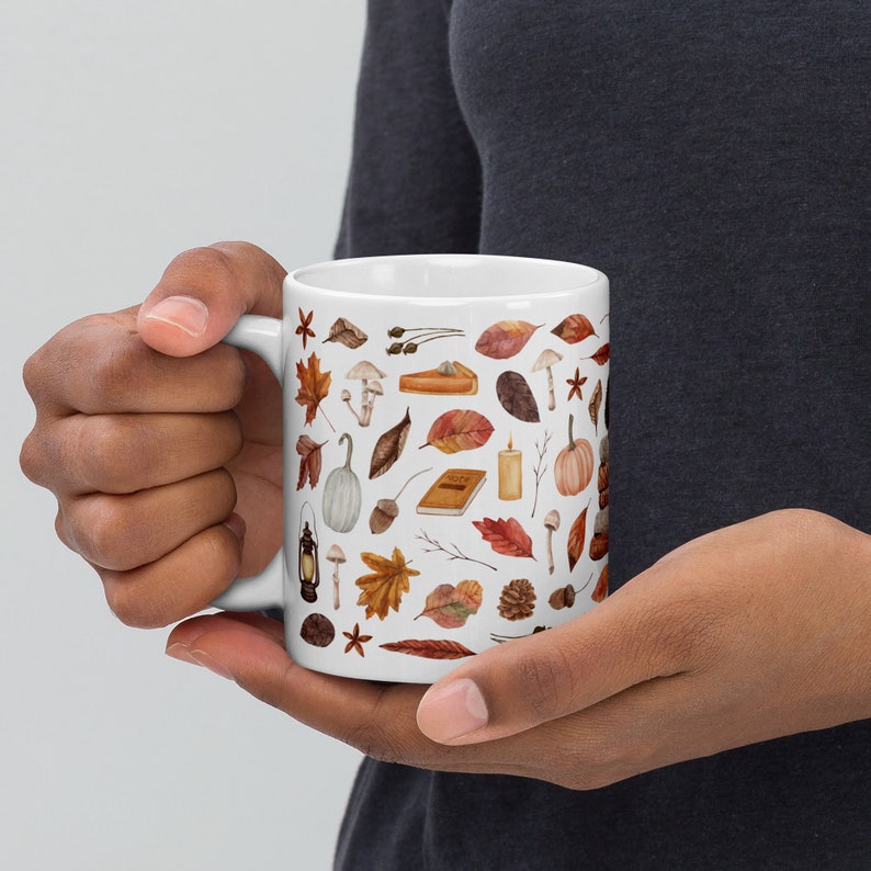 Op de afbeelding: Witte keramische mok met een herfstthema. De mok is bedekt met een patroon van herfstbladeren, pompoenen, eikels, paddenstoelen en andere herfstachtige afbeeldingen.
