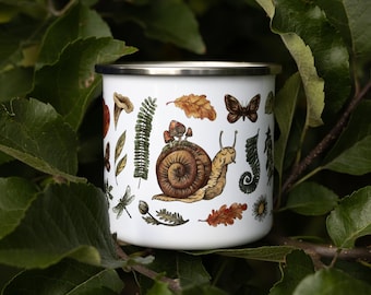 May include: A white enamel mug with a silver rim featuring a detailed illustration of a brown snail with mushrooms on its shell, surrounded by various botanical elements including oak leaves, ferns, a dragonfly, and a butterfly in earthy tones.