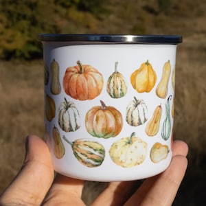 Puede incluir: Una taza de esmalte blanco con un colorido estampado estilo acuarela de varias calabazas y calabacines en tonos naranja, amarillo, verde y crema. La taza tiene un cuerpo cil&iacute;ndrico liso y un borde superior de metal plateado.