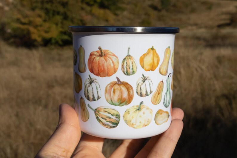 Puede incluir: Una taza de esmalte blanco con un colorido estampado estilo acuarela de varias calabazas y calabacines en tonos naranja, amarillo, verde y crema. La taza tiene un cuerpo cil&iacute;ndrico liso y un borde superior de metal plateado.