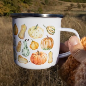 Puede incluir: Una taza de esmalte blanco con borde plateado y asa blanca, que presenta un estampado estilo acuarela de varias calabazas en tonos naranja, amarillo y verde, distribuidas por toda la superficie exterior de la taza.