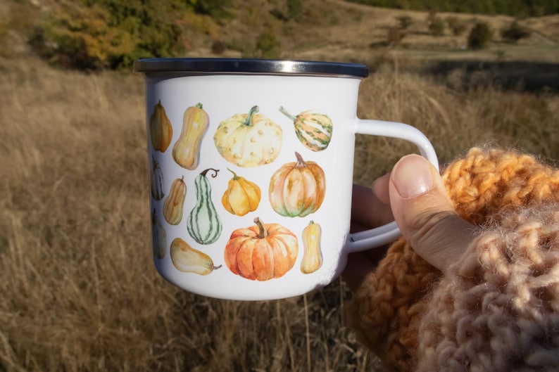 Puede incluir: Una taza de esmalte blanco con borde plateado y asa blanca, que presenta un estampado estilo acuarela de varias calabazas en tonos naranja, amarillo y verde, distribuidas por toda la superficie exterior de la taza.