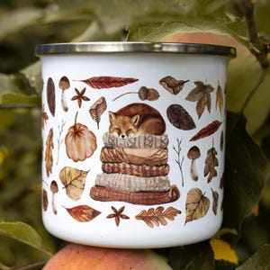 May include: A white enamel mug with a silver rim featuring a watercolor illustration of a sleeping fox curled up on a stack of folded sweaters, surrounded by various autumn leaves, mushrooms, acorns, and star anise in shades of brown and orange.