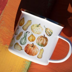 Puede incluir: Una taza de esmalte blanco con un estampado de acuarela colorido de varias calabazas y calabacines en tonos naranja, amarillo y verde. La taza tiene un acabado blanco liso, un borde de metal plateado en la parte superior y un asa lateral.