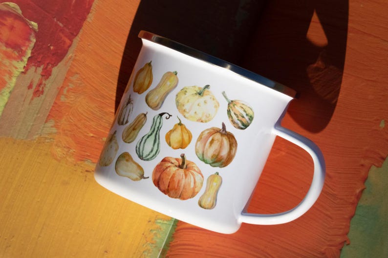 Puede incluir: Una taza de esmalte blanco con un estampado de acuarela colorido de varias calabazas y calabacines en tonos naranja, amarillo y verde. La taza tiene un acabado blanco liso, un borde de metal plateado en la parte superior y un asa lateral.