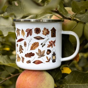 May include: A white enamel mug with a silver rim and a handle, featuring a repeating pattern of brown and orange autumn leaves, acorns, a small wooden birdhouse, and a vintage-style lantern printed on the exterior surface of the cup.